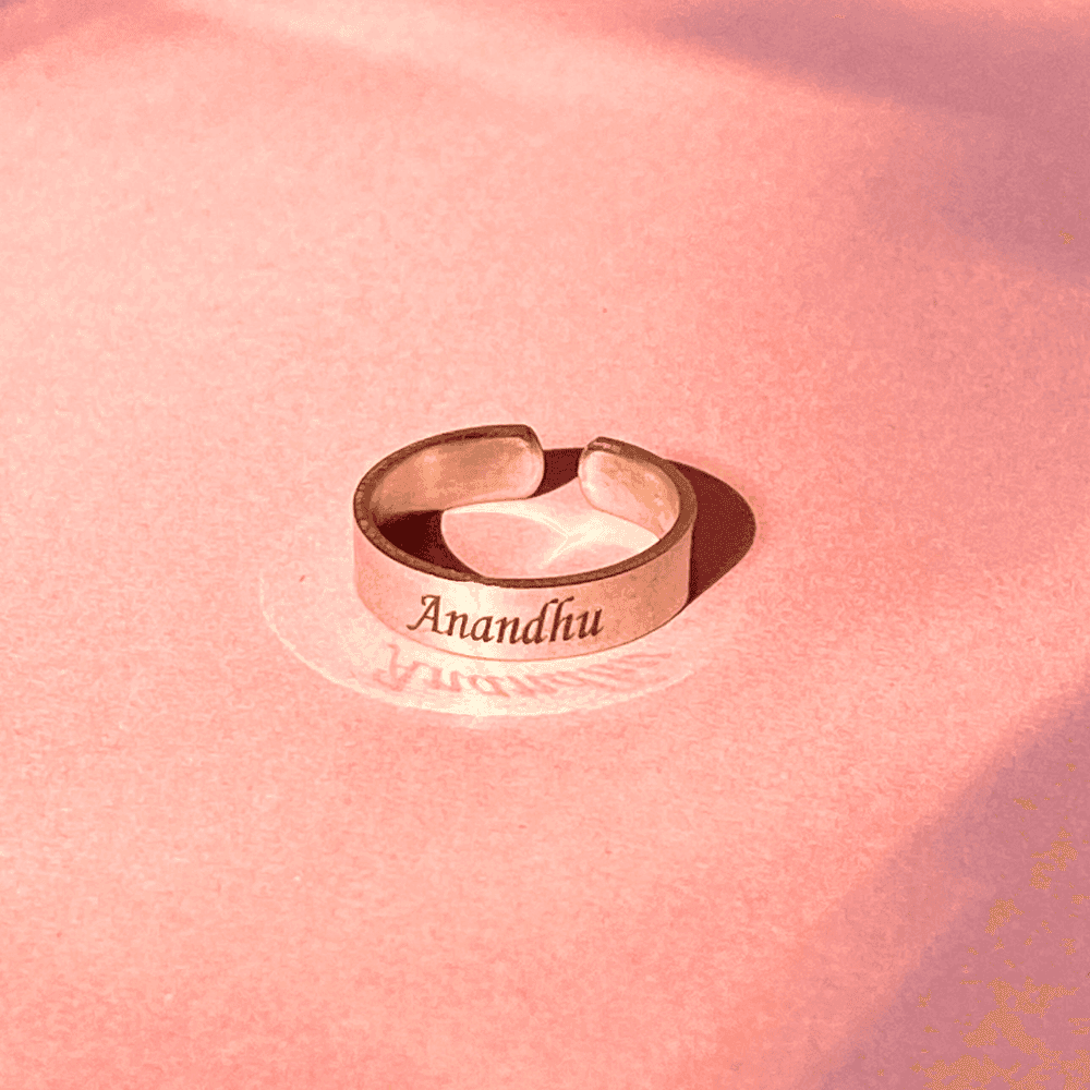 Silver adjustable name ring engraved with Anandhu on a pink background from Ankx.in