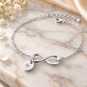 Close-up of a hand wearing a polished silver infinity bracelet with an engraved M charm, styled with a soft cream sleeve and warm floral background.