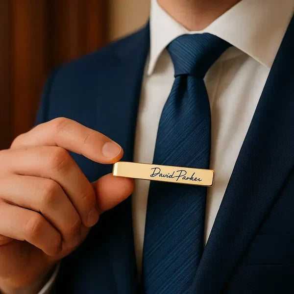 Man in navy suit holding a gold personalized tie clip engraved with "David Parker" near his tie