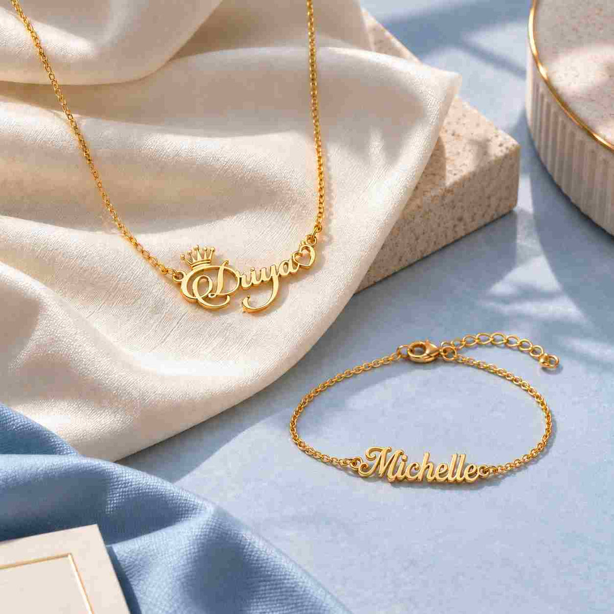 Personalized gold name necklace and name bracelet styled on ivory and soft blue fabric in a premium jewelry setting.