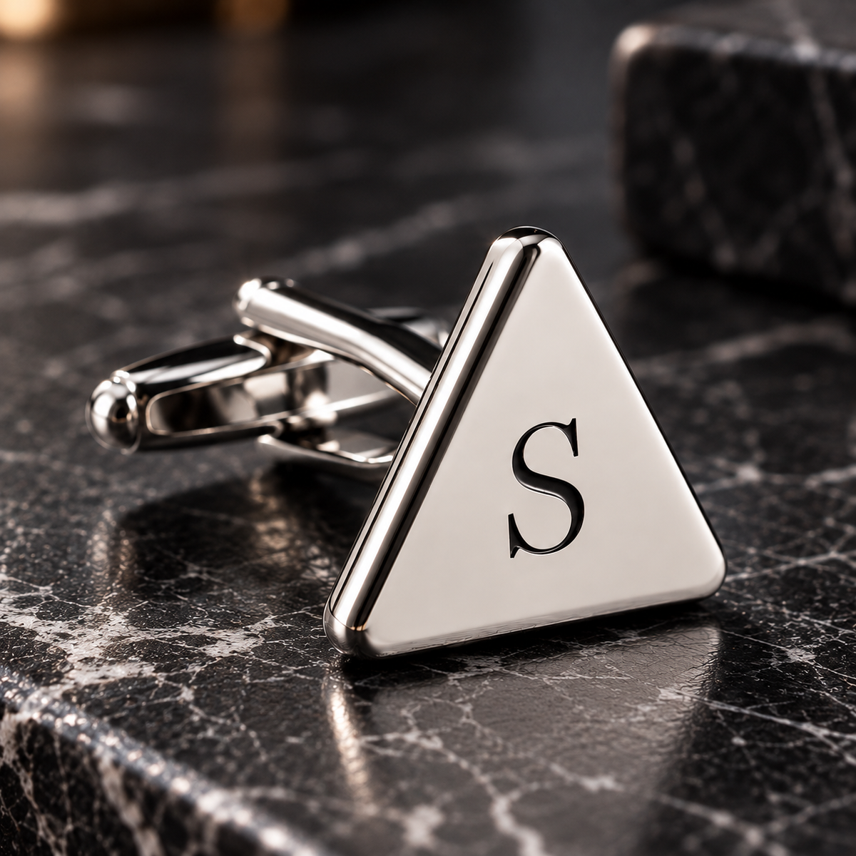 Personalized silver triangular cufflink with black engraved S initial on dark marble surface