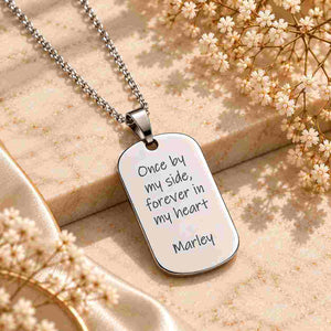 Silver personalized dog tag necklace with “Once by my side, forever in my heart” and “Marley” engraving, displayed on a beige marble surface.
