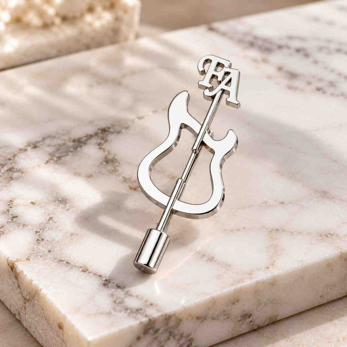 Silver personalized guitar brooch displayed on a light beige marble surface with FA initials