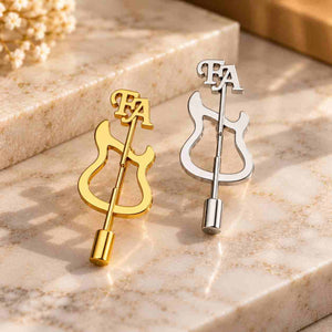 Gold and silver guitar-shaped personalized brooches with FA initials displayed together on a luxury marble surface