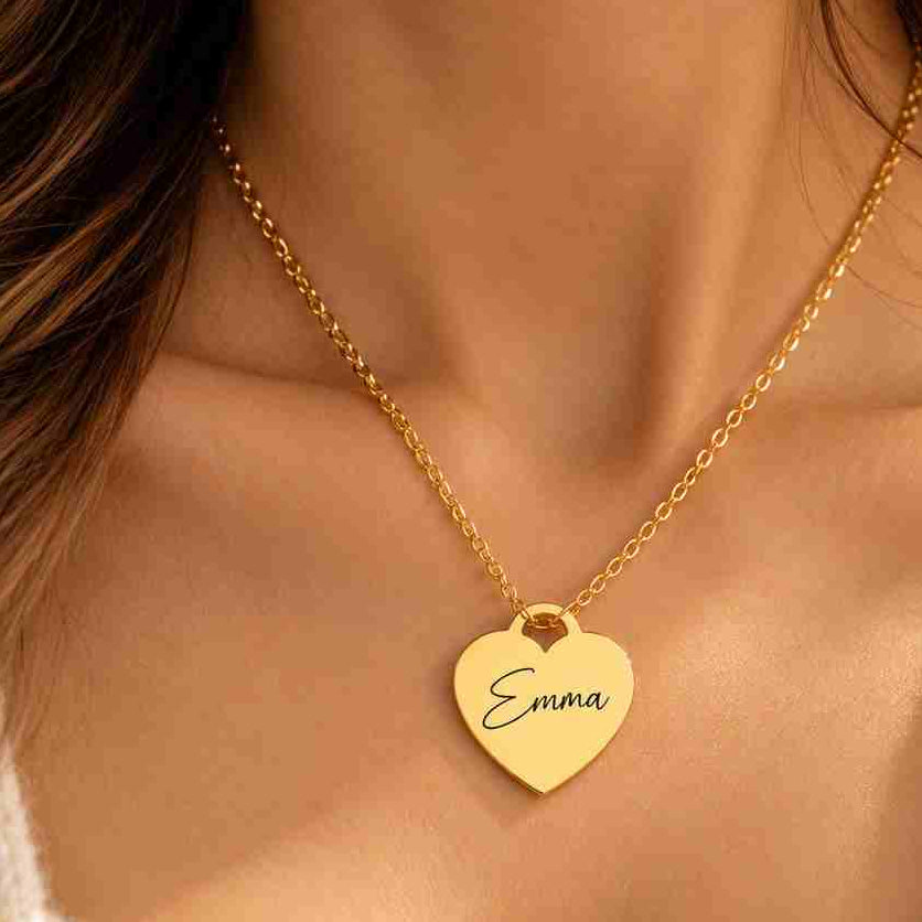 A close-up of a woman wearing a delicate gold chain necklace with a heart-shaped pendant engraved with the name 'Emma'.