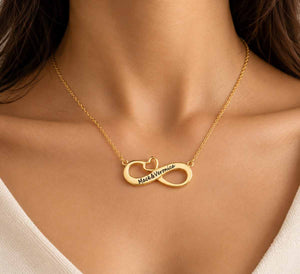 A close-up of a gold infinity necklace worn on a model's neck. The pendant features one loop engraved with the names 'Jack & Niamh' in black script, and the other loop contains a small heart-shaped cutout.