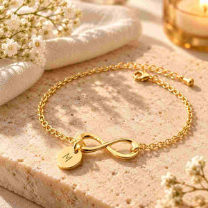 Close-up of a hand wearing a delicate gold infinity bracelet with an engraved M round charm, styled with a cream sleeve and soft white flowers.
