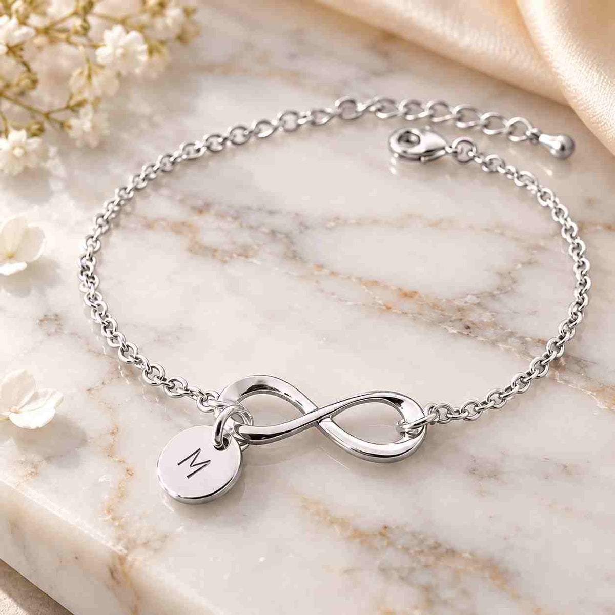Close-up of a hand wearing a polished silver infinity bracelet with an engraved M charm, styled with a soft cream sleeve and warm floral background.