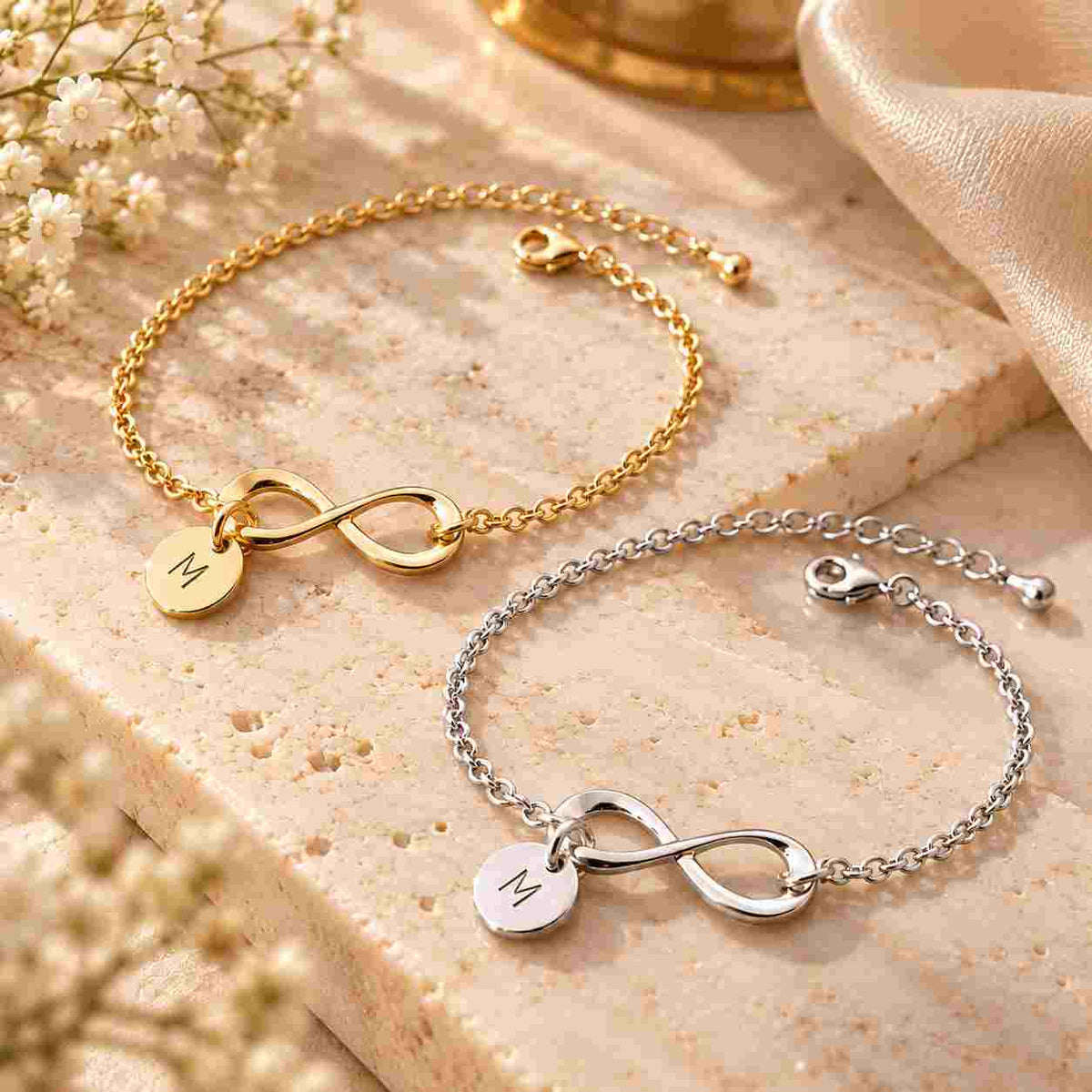 Gold and silver personalized infinity bracelets with engraved M charms displayed together on a warm beige stone surface in an elegant jewellery setting.