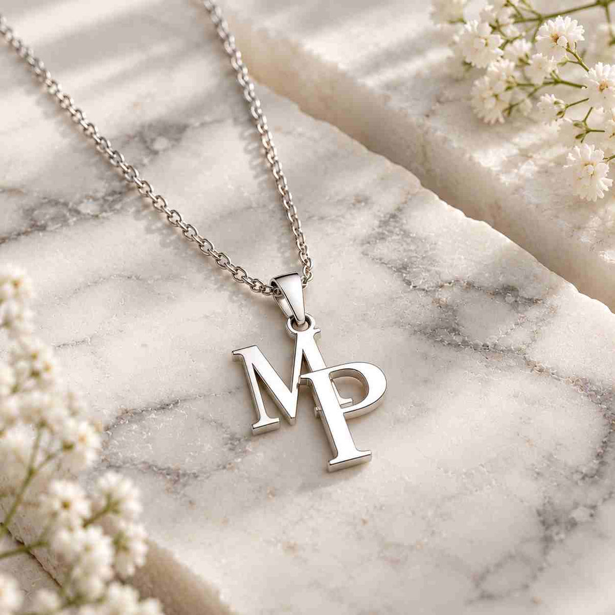 A silver initial necklace with the letters 'M' and 'P' in a monogram pendant, displayed on a white marble slab with small white flowers.