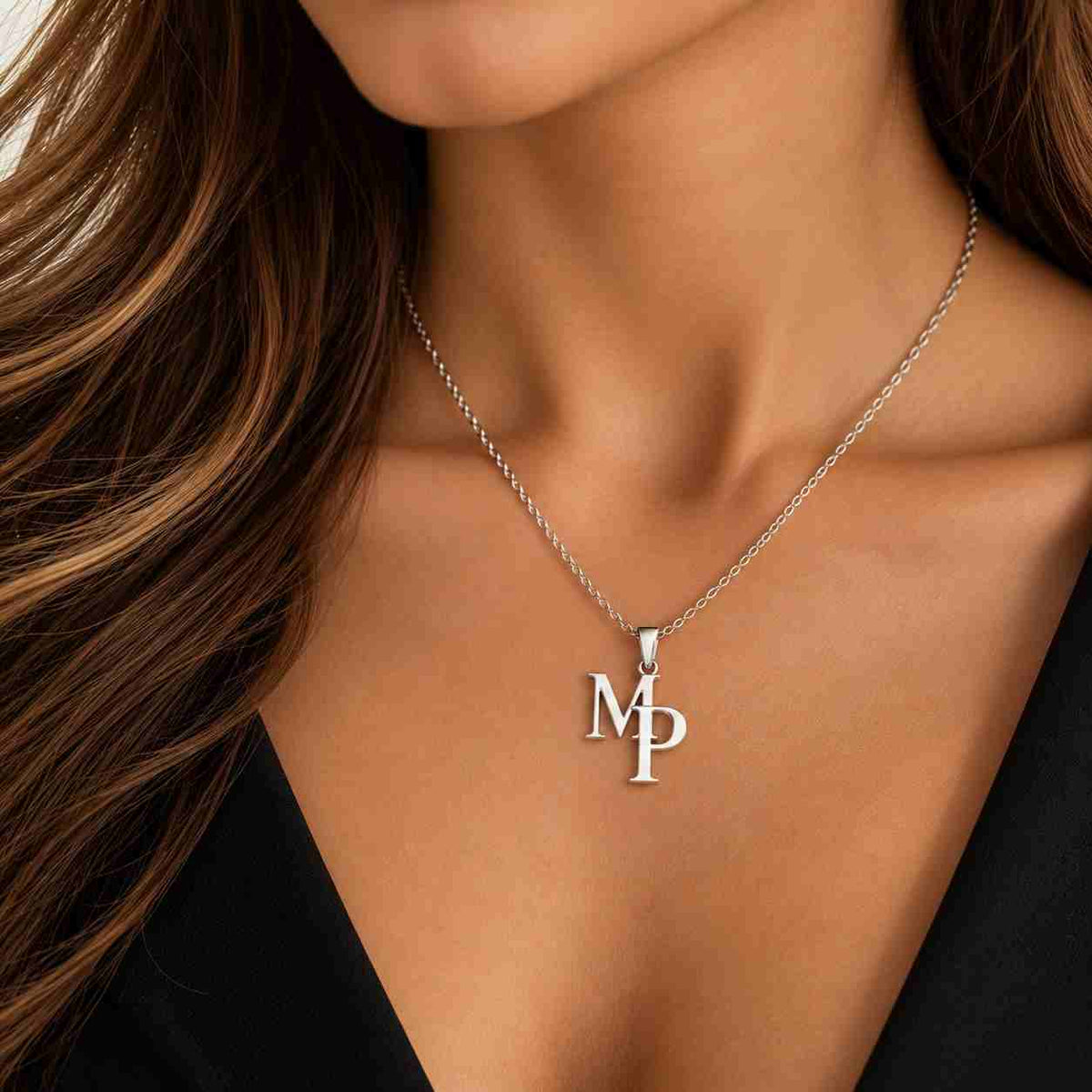 A close-up of a woman wearing a delicate silver chain necklace with an interlocking 'MP' initial pendant, styled with a black top.